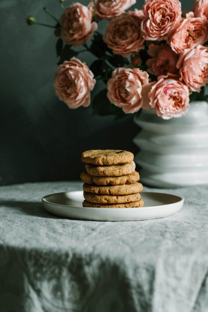 Delicious homemade cookies stacked on a plate with pink roses in a vase beside them.